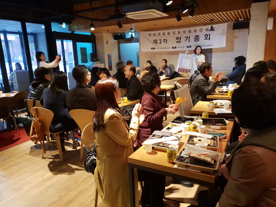 정기총회 현장, 발표자가 마이크를 들고 인사말을 하는 모습과 박수치는 참석자들