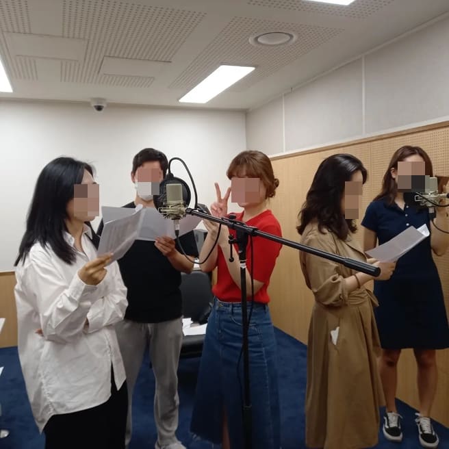 녹음 부스에서 여러 명이 원고를 들고 마이크 앞에 서 있는 모습(낭독 녹음)